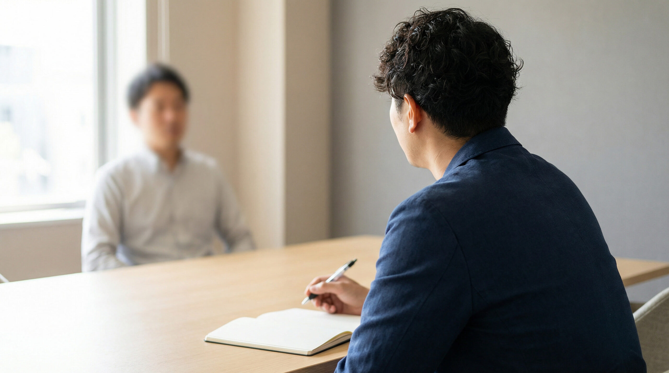 A hiring manager sits across from a job candidate in a bright office, taking notes during a behavioral interview. The scene shows a simple wooden table, natural light, and a neutral professional environment.