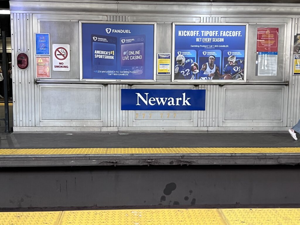 Newark PATH train station platform with sports betting advertisements and a bold blue Newark sign, capturing a quiet moment before heading into New York City — a scene that sets the tone for reflecting on bar exam mindset and personal growth.